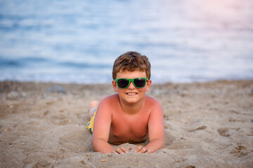 Little boy kid with sunglasses is lying on the sand near seaside smiling after sunburn.