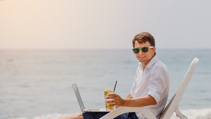 Young man in sunglasses and white shirt is lying on a beach chair on the seaside holding a cocktail and a laptop.