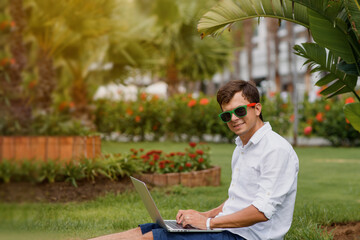 Young man in sunglasses and white shirt is lying on grass  and working in a tropical location