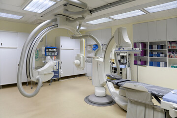 A modern hospital room, a large portable mobile scanning machine with curved shaped arms, a mobile scanner and hospital gurney or bed.