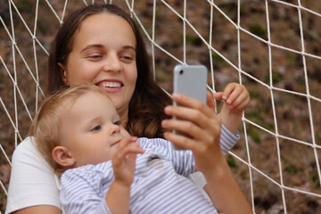 Portrait of young smiling attractive woman with her daughter, mother and child relaxing in hammock in forest and using smart phone, looking at device screen, watching online cartoons together.