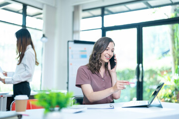 Fototapeta premium Young business woman on the phone at the office.Business woman texting on the phone and working on laptop.Beautiful young business woman sitting in office.Business woman smiling.