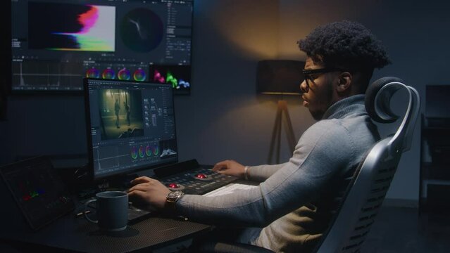 African American film maker makes color grading on computer in modern studio. Big screen with RGB colour correction graphic bar on wall. Action film footage displayed on monitor. Colorist equipment.