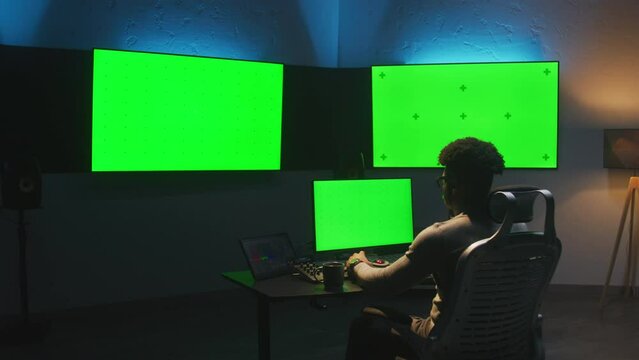 African American man works on computer in studio with equipment. Multiple chroma key big screen on the wall. Professional software displayed on tablet. Color grading or film editing room. Dolly shot.