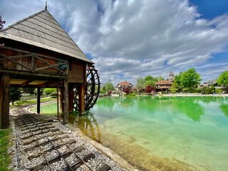 Scenic view of a traditional wooden watermill with a large water wheel next to a turquoise lake. Bright summer sky with clouds reflecting in the water creates a picturesque atmosphere. Perfect represe