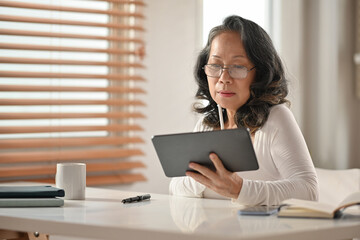Senior Asian woman wearing glasses reading online news or shopping online on digital tablet 