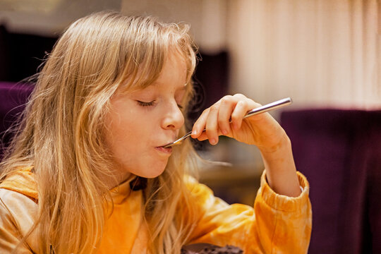 little girl with loose white hair happily eats a cake with a fork in a cafe. horizontal