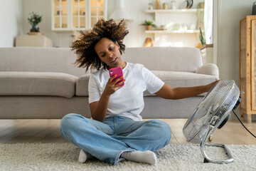 African American girl sitting on floor in living room near electric fan and using smartphone,...