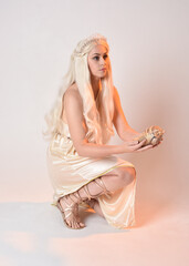 Full length portrait of beautiful blonde woman wearing a fantasy goddess toga costume with  magical crown.
Seated pose holding an ocean seashell, sitting on floor. isolated on white studio background.