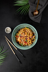 Chinese fried noodles with chicken and vegetables in ceramic bowl on black concrete background....