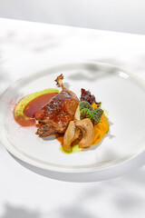 Traditional france dish - duck confit with baked apple on light background with shadows of sunlight. Roasted duck leg with fruits and gravy on white plate in summer dining Elegant roasted leg
