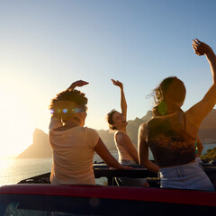 Female Friends Standing Up Through Sun Roof Car And Dancing On Road Trip Through Countryside