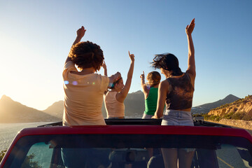 Female Friends Standing Up Through Sun Roof Car And Dancing On Road Trip Through Countryside