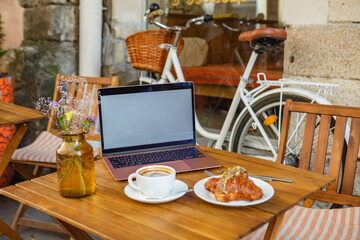 White laptop screen, croissant and coffee on the summer terrace of a cafe