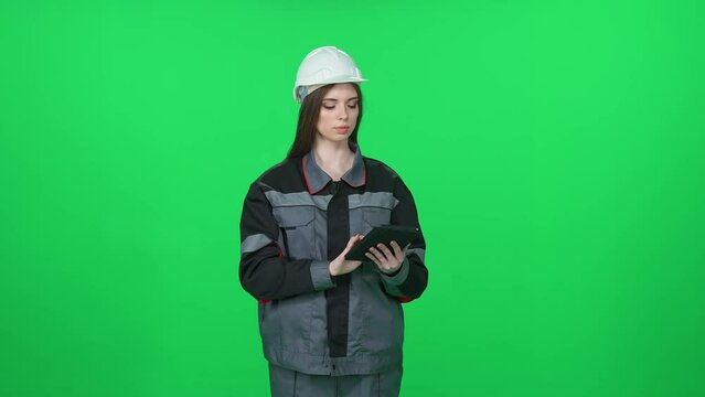 Digital engineer, woman worker in a uniform and protective helmet, technical engineer stands and uses a screen tablet on a green background, woman engineer on chromakey.