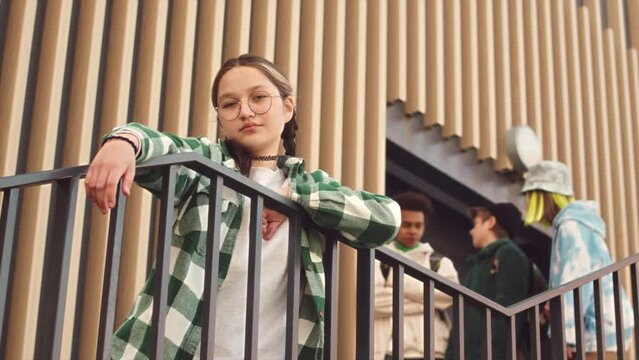 Low angle slowmo portrait of cool 14 year old Caucasian girl in checkered shirt and round eyeglasses posing for camera outdoors in summer while her friends chatting in background