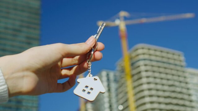 Woman's hand holds key and keychain in form of house against background of house under construction. concept of own housing. Buying apartment from real estate agency mortgage. Housewarming settlement