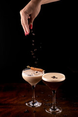 female hand pouring roasted coffee beans into espresso martini cocktail and baileys in fluted glasses and cinnamon stick close-up