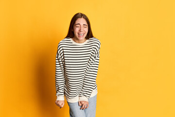 Obraz premium Portrait of young beautiful girl in striped sweater smiling, laughing against yellow studio background. Funny jokes and good mood. Concept of youth, emotions, facial expression, lifestyle