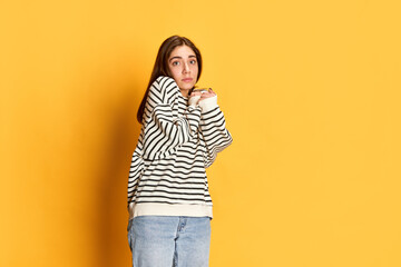 Studio portrait of young girl in stripes sweater posing with little fear in eyes agaisnt yellow...