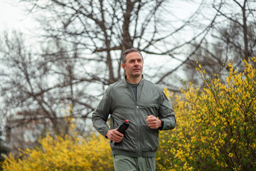 A middle shot of a fit middle-aged man jogging in a city park with a yellow flower bush in the...