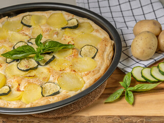 A pan of quiche with potatoes and sliced zucchini on a wooden cutting board with whole potatoes beside it