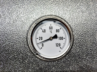 Industrial circular arrow-pointer display for temperature readings in degrees Celsius is mounted on a shiny textured metal surface. The arrow indicates the value ten.