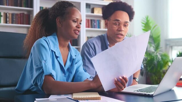 Young professional colleagues Arabian man and African American woman doing paperwork together looking through company financial records dressed in casual style sits at table in home office