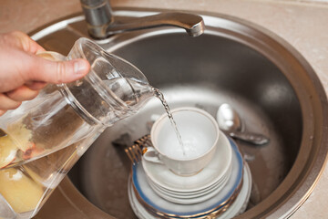 A man with a decanter in his hand pours water onto dishes, forks, spoons, plates, cups, in the washbasin. Kitchen care, apartment cleaning, cleanliness.