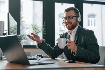 In headphones. Having online conversation. Businessman in suit is in the office with computer