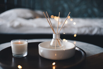 Liquid home fragrance in glass bottle with bamboo sticks and scented candle on wooden tray on table over bed and glowing lights at night close up. Cozy hygge atmosphere.