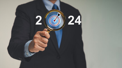 Businessman holding a magnifier with a darts board icon and the letters 2024 while standing against...