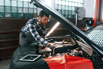 Under the hood. Holding lighting equipment and fixing the issues of the car. Man in uniform is working in the auto salon