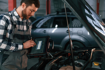 Standing near the car with hood opened. Holding tablet. Man in uniform is working in the auto salon