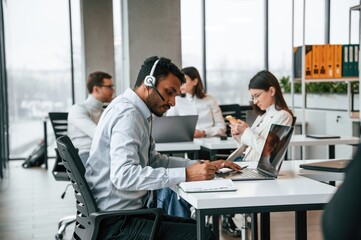 Man is sitting by laptop in headphones. Four people are working in the office together