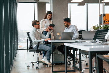 By the table with laptop. Men and woman are working in the office together