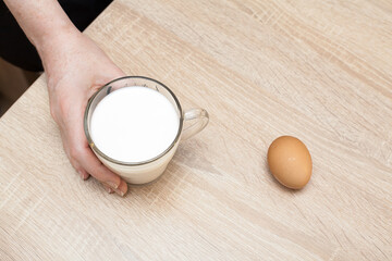 A woman's hand holds a glass of fresh milk on a textured table, a brown egg lies nearby. An image for your healthy food creatives.