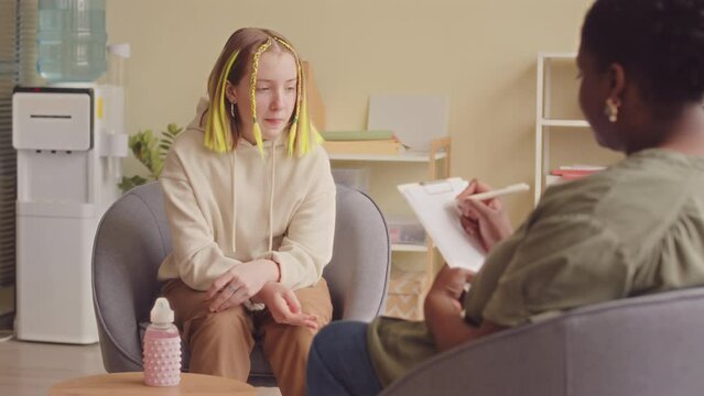 Medium slowmo of 14 year old Caucasian teenage girl with dyed yellow hair speaking about her mental state with Black female psychologist during individual therapy session