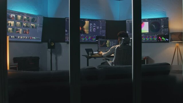 Professional African American video editor makes color grading in program on PC. Process of colour correction for movie post production in modern studio. Multiple monitors with action film footage.