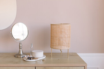 Rattan lamp, mirror, feminine accessories on ceramic plate on wood console in bedroom. Copy space. 