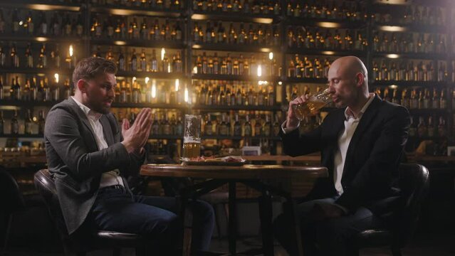 Two friends are relaxing in a pub after work on Friday. The men, dressed in jackets and shirts, are drinking beer. A man offers his colleague a drink, but the colleague refuses.