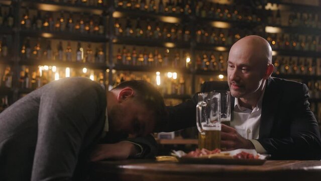 Two friends are relaxing in a pub after work. The men, dressed in jackets and shirts, are drinking beer. A person is drunk and has fallen asleep at the table. Colleague is trying to wake them up.