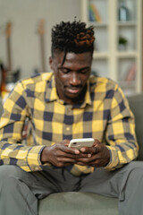 Handsome African man using smartphone while sitting on a sofa in cozy living room. Young man typing a message