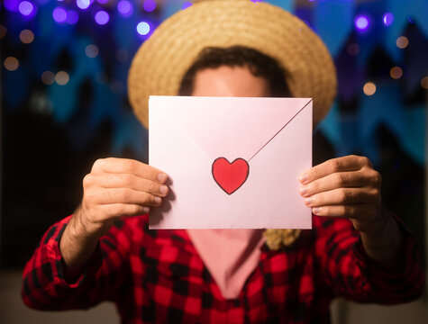 Brazilian June Festival, close up of valentine card with heart shape, known as Correio Elegante, in the hand of anonymous man with red plaid shirt and straw hat at night.