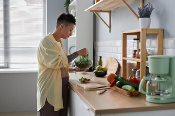 Side view portrait of pregnant young woman cooking healthy meal in kitchen and making salad with green herbs