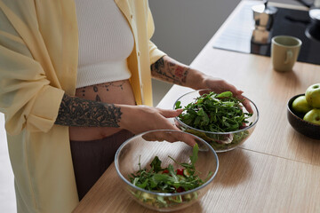Close up of pregnant young woman with tattoos making salad in kitchen and holding bowl of green herbs