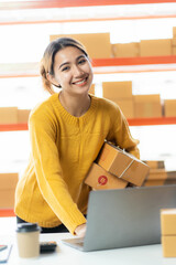 Asian businesswoman working with yellow boxes to start online delivery. Freelancer or salesperson checking production orders with laptop. SME entrepreneur making parcel boxes at home.