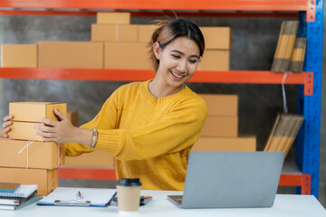 Asian businesswoman working with yellow boxes to start online delivery. Freelancer or salesperson checking production orders with laptop. SME entrepreneur making parcel boxes at home.