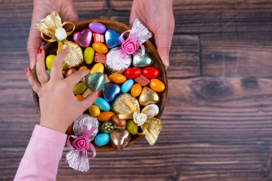 Serving confectionery, top view image of woman hand holding serving confectionery. Child girl taking one of the sweets. Wrapped chocolates, almond dragee. Wooden background, copy space.
