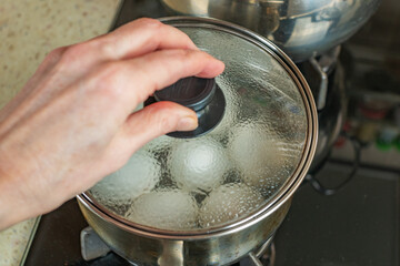 Pot of boiling water in which white chicken eggs are boiled. Water boils and boils in pan with boiled eggs. Female hand closes pan with boiled white chicken eggs with transparent glass roof. Close-up